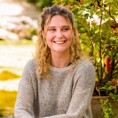Young woman with blonde curly hair sitting on a rock in a garden wearing dark trousers and grey knitted jumper, arms lightly folded.